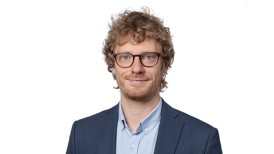 A man with curly hair, glasses, wearing a light blue shirt and dark blue jacket.