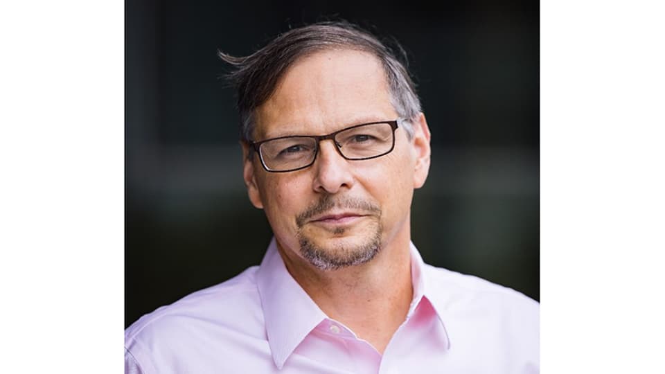 A dark-haired man in glasses, wearing a pink shirt.