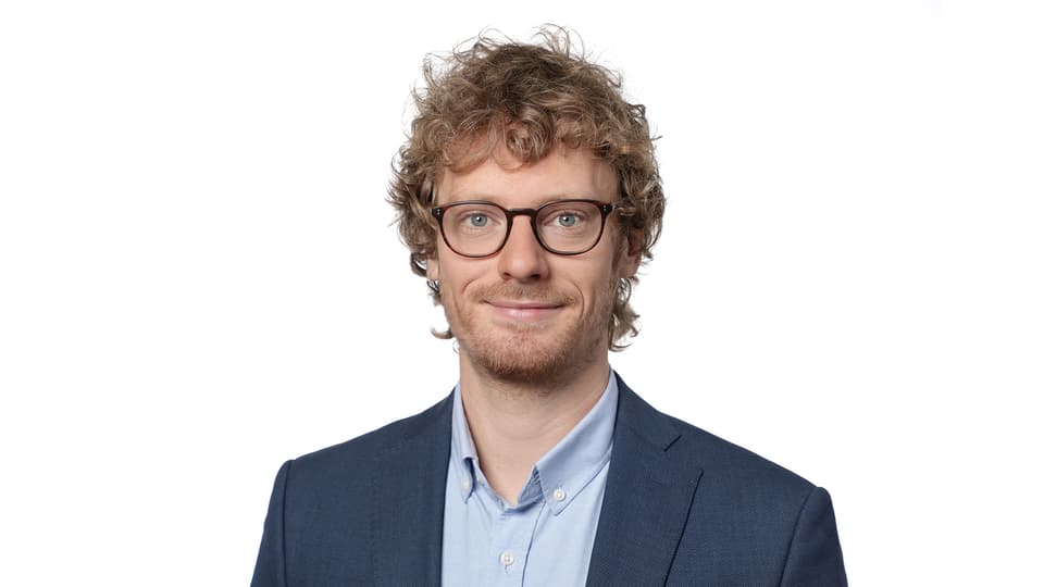 A man with curly hair, glasses, wearing a light blue shirt and dark blue jacket.